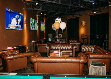 Cozy sports lounge interior with tufted brown leather sofas arranged around a coffee table topped with gold and white balloons, wall-mounted TVs showing sports, warm lighting and a pool table in the foreground.