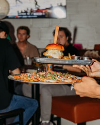 Server carrying a metal tray with a cheeseburger skewered by a wooden knife, fries and dipping cups, plus a plate of loaded nachos, in a lively casual bar-restaurant setting.