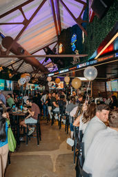 Lively indoor sports bar with exposed wooden beams, neon signs and TV screens, crowded high-top tables and bar stools, gold and white balloons and patrons enjoying drinks.