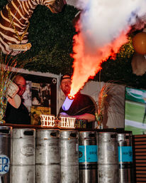 Lively indoor bar scene with metal beer kegs as a counter, person firing a fog cannon that shoots bright orange smoke, glowing "YEE HAW" sign, zebra head decor and festive streamers.