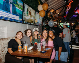 Five smiling friends gathered at a wooden table in a lively sports bar, drinks and beer cans on the table, gold and white balloons overhead, multiple TV screens on the wall creating a festive nightlife vibe.
