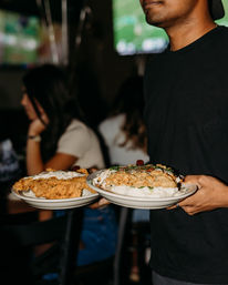 Server carrying two plates of crispy fried chicken on mashed potatoes with gravy in a busy sports bar — hearty comfort-food dinner scene.