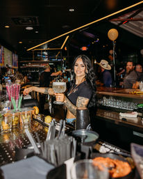 Smiling tattooed bartender with long dark hair holding a large frosted margarita behind a busy bar with garnishes, straws, balloons and nighttime crowd, lively nightlife scene
