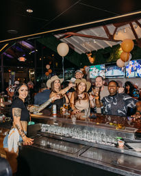 Lively bar scene with friends and a smiling bartender toasting drinks at a packed sports bar — cowboy hats, balloons, TV screens, and nightlife energy.