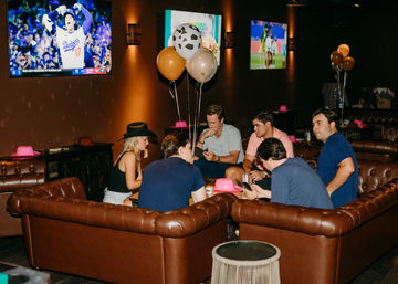 Six friends gathered on brown leather couches in a dim sports lounge with party balloons, pink cowboy hats and TVs showing baseball and soccer