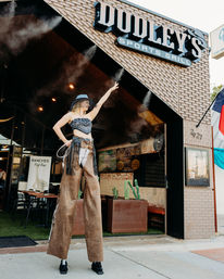 Stilt-walker dressed as a cowgirl—wide-brim hat, bandana top and fringe chaps—striking a pose outside a brick sports-grill entrance with cactus planters and outdoor patio seating.