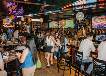 Busy urban sports bar interior with balloons, bar stools and tables, groups chatting and watching football on multiple TVs — lively downtown nightlife scene.