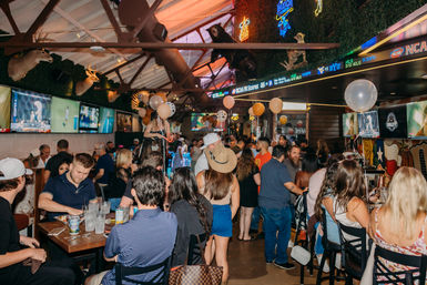 Bustling indoor sports bar with crowds socializing, celebratory balloons, neon signs and multiple TVs showing games, rustic deer-head decor and patrons in casual summer outfits including a cowboy hat.