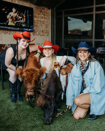 Three smiling women in cowboy hats and boots pose with two small ponies on artificial turf at a western-themed patio