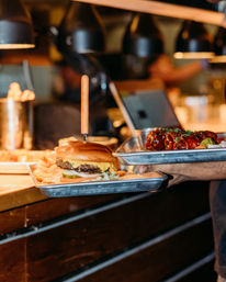Juicy cheeseburger with melted cheddar, pickles and fries on a metal tray skewered with a wooden knife, alongside a tray of glazed BBQ ribs being carried through a restaurant pass under heat lamps.
