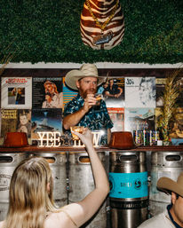 Cowboy-hat bartender offers a cheers with a shot in a lively Western-themed country bar, framed by stacked beer kegs and a vintage vinyl record wall.