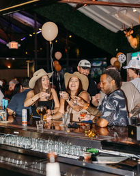 Smiling friends in cowboy hats clinking shot glasses at a lively country-themed bar with balloons, neon lights, and a crowded bar counter — nightlife celebration.
