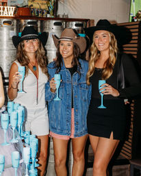 Three friends in cowgirl hats holding turquoise champagne flutes at a bar with stacked kegs — denim jacket, smiles, Western night out.