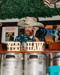 Bearded man in a cowboy hat and patterned western shirt holding headphones behind a bar, neon “YEE HAW” sign above beer kegs and vintage vinyl covers in a lively country-western bar.