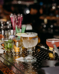 Frosty salt-rimmed margarita goblet with a lime wedge on a metal bar counter, pink cocktail coupe and blurred bar tools in the background, lively cocktail bar scene.