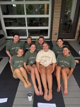 Bachelorette party group photo of nine women, bride in a light tee centered, all smiling on yoga mats on a wooden deck patio in matching shirts.