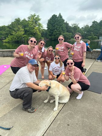 Smiling group of women in matching pink "Let's Go Girls!" shirts and heart-shaped sunglasses holding orange mimosas at a lakefront park, with a man kneeling to pet a yellow Labrador and yoga mats on the pavement.