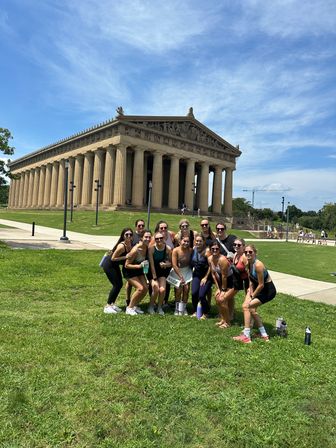 Group of women in sporty outfits posing on a grassy lawn in front of the Parthenon replica at Centennial Park in Nashville under a bright blue sky