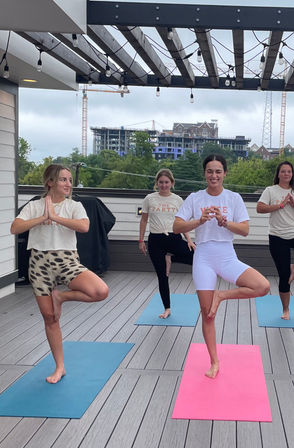 Outdoor rooftop yoga class on a wood deck with string lights, four women smiling in tree pose on colorful mats with urban skyline and construction cranes in the background.