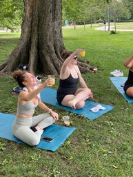Three women seated on blue yoga mats under a large tree in a park, laughing and toasting with orange drinks after outdoor yoga