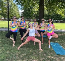 Six women in tie-dye tanks and sunglasses practicing yoga in a sunny city park, holding tree pose on the grass near a large shade tree with yoga mats nearby.