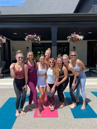 Eight women smiling on colorful yoga mats during an outdoor yoga session on a sunny backyard patio with hanging flower baskets