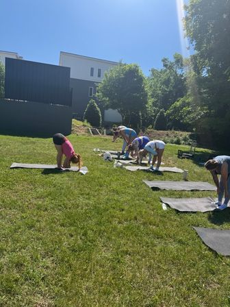 Group outdoor yoga on a sunny neighborhood lawn — people bending forward on mats under a clear blue sky with trees and modern houses in the background.