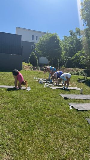 Group outdoor yoga on a sunny neighborhood lawn — people bending forward on mats under a clear blue sky with trees and modern houses in the background.