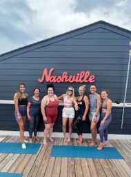Cheerful group of seven women in activewear posing barefoot on a wooden rooftop deck with blue yoga mats beneath a red Nashville sign on dark siding and a cloudy sky above