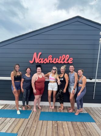 Cheerful group of seven women in activewear posing barefoot on a wooden rooftop deck with blue yoga mats beneath a red Nashville sign on dark siding and a cloudy sky above