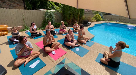 Group of women practicing guided outdoor yoga and meditation on colorful mats around a suburban backyard swimming pool under a sun umbrella