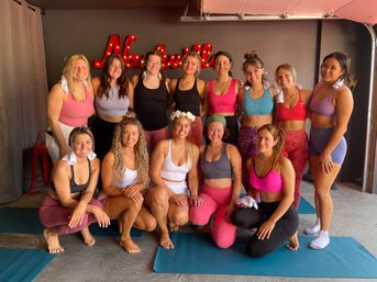 Smiling group of women in colorful activewear posing on yoga mats in a bright Nashville fitness studio with a red neon sign, post-yoga class vibes.