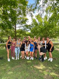 Smiling group of women in colorful activewear posing under leafy trees by a pond in a sunny park — outdoor fitness meetup and friends portrait.