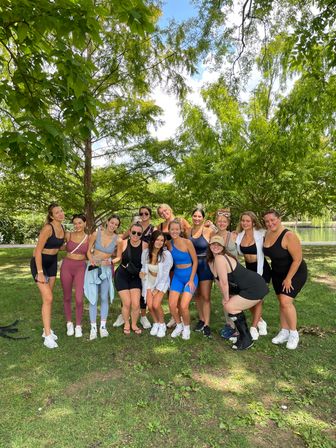 Smiling group of women in colorful activewear posing under leafy trees by a pond in a sunny park — outdoor fitness meetup and friends portrait.