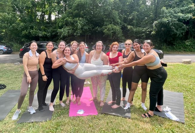 Smiling group of women holding a friend horizontally during an outdoor yoga session on mats in a leafy park