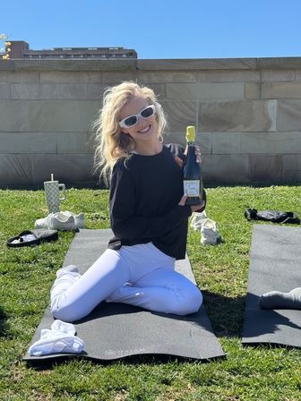 Smiling person on a yoga mat during outdoor yoga on a sunny grassy lawn, wearing white sunglasses and leggings and holding a champagne bottle.