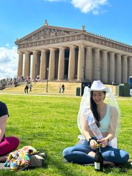 Bride-to-be wearing a white cowboy hat, veil and sash pops a champagne bottle while sitting on the green lawn in front of the Parthenon replica at Nashville’s Centennial Park on a sunny day.