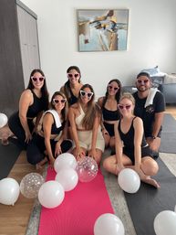 Seven friends wearing heart-shaped sunglasses smiling on yoga mats surrounded by white balloons in a bright apartment living room