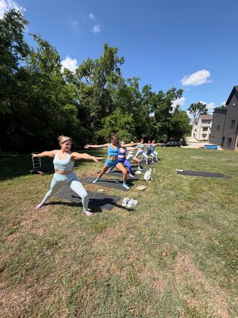 Sunlit outdoor yoga class on a grassy suburban lawn — women in colorful activewear holding Warrior II on mats with trees and townhomes in the background.