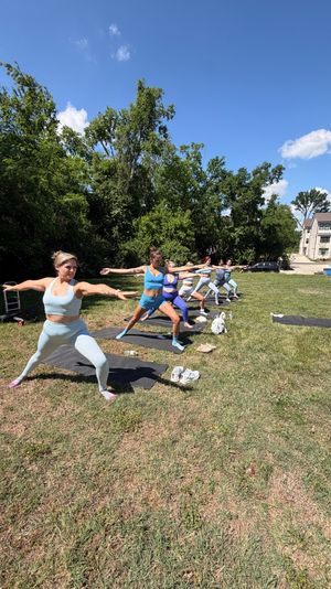 Sunlit outdoor yoga class on a grassy suburban lawn — women in colorful activewear holding Warrior II on mats with trees and townhomes in the background.