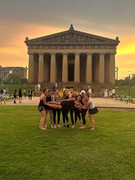 Group of friends playfully lifting a person on the grassy lawn in front of the Parthenon replica at Centennial Park, Nashville, Tennessee, illuminated by a golden sunset