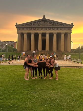 Group of friends playfully lifting a person on the grassy lawn in front of the Parthenon replica at Centennial Park, Nashville, Tennessee, illuminated by a golden sunset