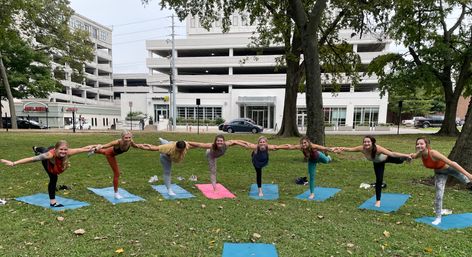 Eight people balancing in a connected yoga pose on colorful mats in a grassy urban park, trees framing a multi-level parking garage and buildings in the background.