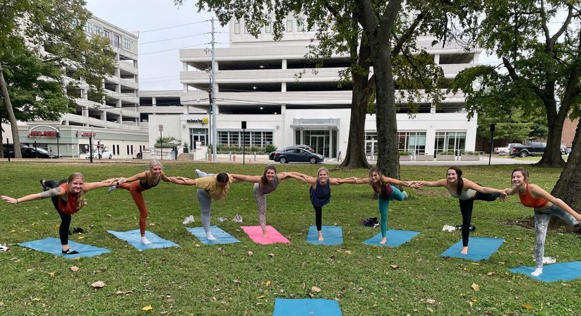 Eight people balancing in a connected yoga pose on colorful mats in a grassy urban park, trees framing a multi-level parking garage and buildings in the background.