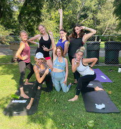 Seven women in activewear posing playfully on yoga mats during an outdoor group fitness/yoga class on a sunny park lawn with trees