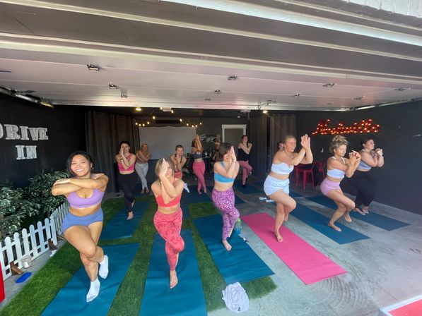 Upbeat group yoga class in a converted garage studio: women on colorful mats practicing single-leg balance poses under string lights.