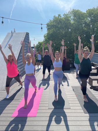 Group rooftop yoga class on a sunny outdoor deck, participants in high lunge with arms raised on colorful mats, string lights overhead and leafy trees in the background.