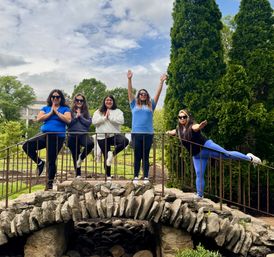 Five friends striking outdoor yoga and balance poses on a rustic stone footbridge in a leafy public garden with tall evergreens and a cloudy blue sky