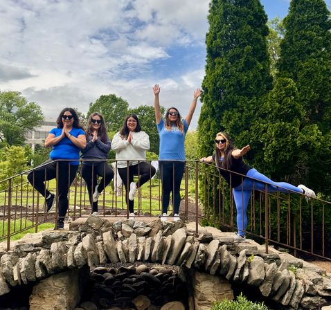 Five friends striking outdoor yoga and balance poses on a rustic stone footbridge in a leafy public garden with tall evergreens and a cloudy blue sky