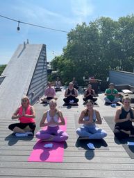Sunlit rooftop yoga class on an urban terrace with string lights, participants seated in lotus pose with hands in prayer on colorful mats, trees and blue sky in the background.